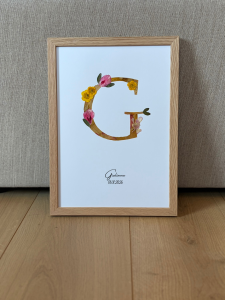 composition florale d'un monogramme réalisé à la main, entièrement composée de fleurs préssées. Ce monogramme représente la lettre G du prénom Giulianna. Est annoté en dessous le nom est la date de naissance