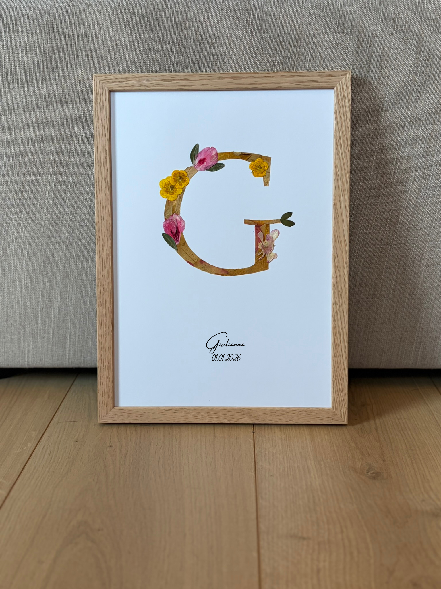 composition florale d'un monogramme réalisé à la main, entièrement composée de fleurs préssées. Ce monogramme représente la lettre G du prénom Giulianna. Est annoté en dessous le nom est la date de naissance
