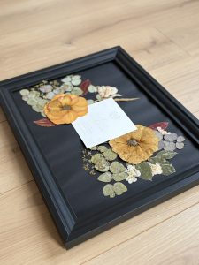Une composition florale élégante réalisée à la main à partir de fleurs et feuillages soigneusement pressés. De grandes fleurs aux teintes chaudes d’ocre et de jaune s’harmonisent avec l’eucalyptus et de délicates touches végétales, créant un équilibre raffiné sur un fond noir profond. Au centre, le faire-part personnalisé est sublimé par l’arrangement floral, comme suspendu dans un écrin naturel. Encadrée dans un cadre noir intemporel, cette création transforme un souvenir précieux en œuvre décorative unique, à conserver pour toujours.