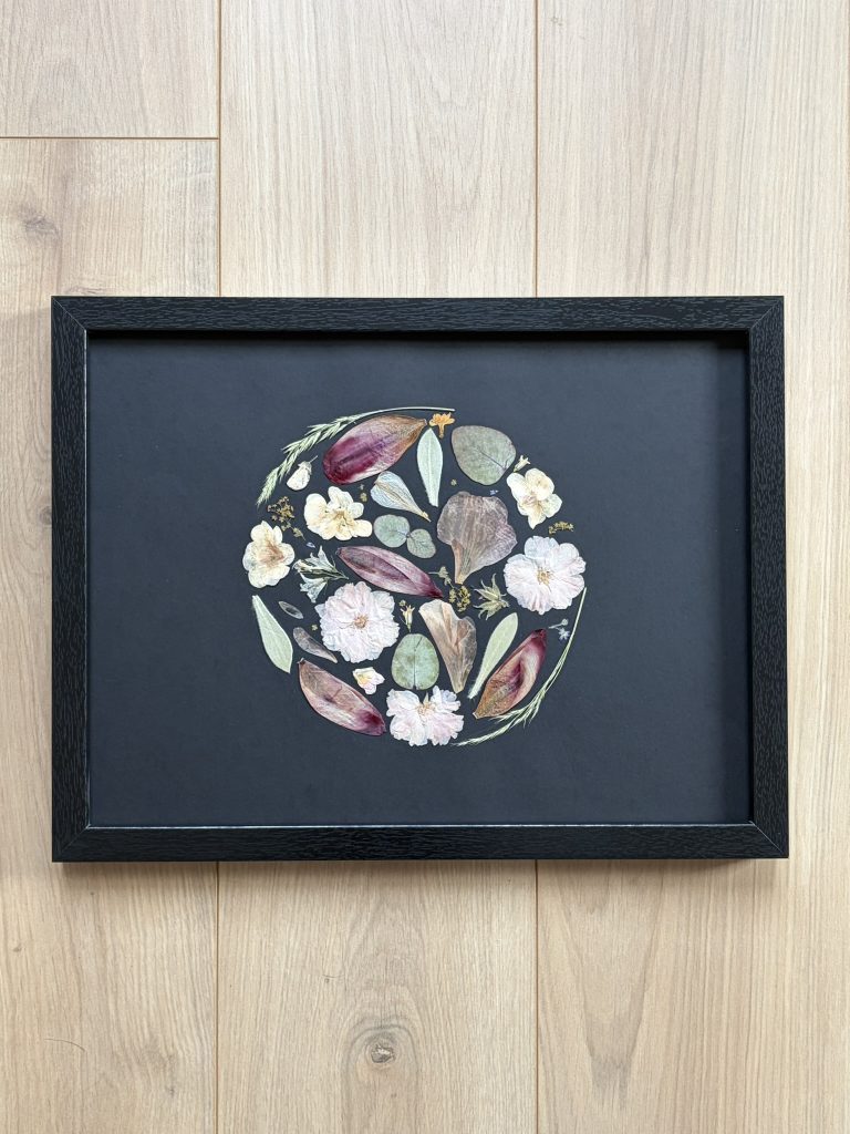 Une composition florale circulaire réalisée à la main à partir de fleurs et feuillages délicatement pressés. Les nuances douces de rose poudré, de bordeaux et de vert eucalyptus se déploient harmonieusement sur un fond noir profond, créant un contraste élégant et contemporain. Chaque élément est minutieusement disposé pour former un équilibre naturel et raffiné. Encadrée dans un cadre noir intemporel, cette création devient une pièce décorative unique, à la fois artistique et poétique, qui sublime la beauté fragile des fleurs préservées.