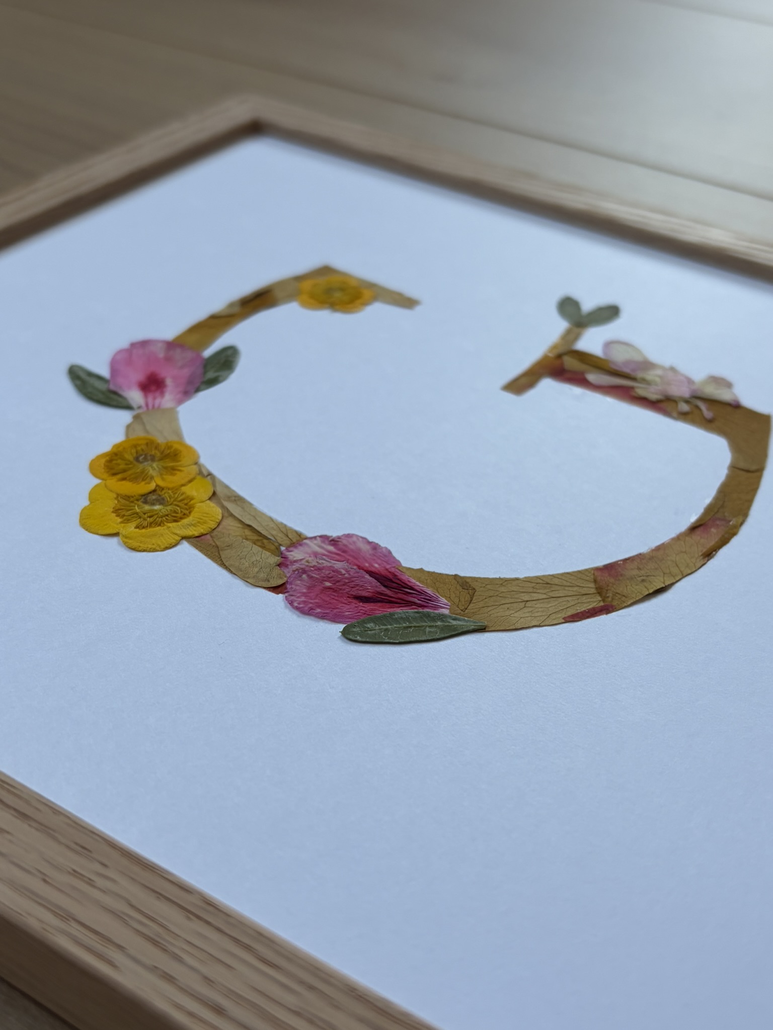 n monogramme floral réalisé à la main à partir de véritables fleurs pressées. La lettre G, délicatement composée de pétales roses et jaunes, est encadrée de bois naturel pour une finition douce et épurée. Personnalisé avec le prénom Giulianna et sa date de naissance, ce cadre devient un souvenir unique et intemporel, parfait pour célébrer une naissance avec poésie.